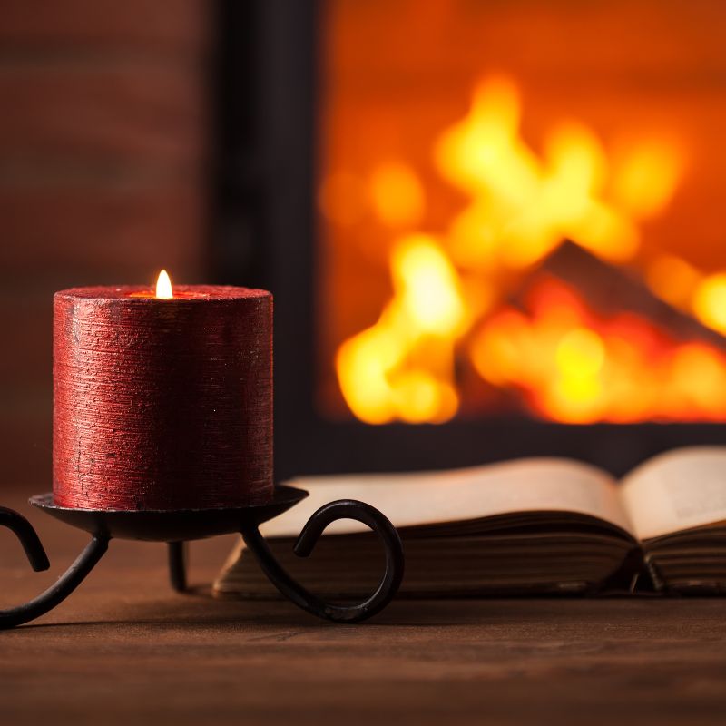a red lit candle on a little candle stand next to an open book with a lit fireplace in the background